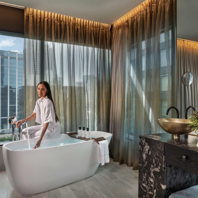 A woman in a white robe sits on a freestanding bathtub in a modern bathroom with large windows and sheer curtains. The mood is serene and luxurious.