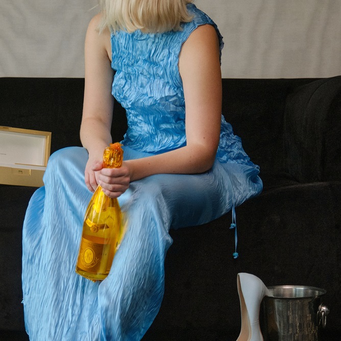 A person in a blue dress holds a yellow champagne bottle, sitting on a dark sofa. A shoebox and high heel are nearby, suggesting a celebratory mood.