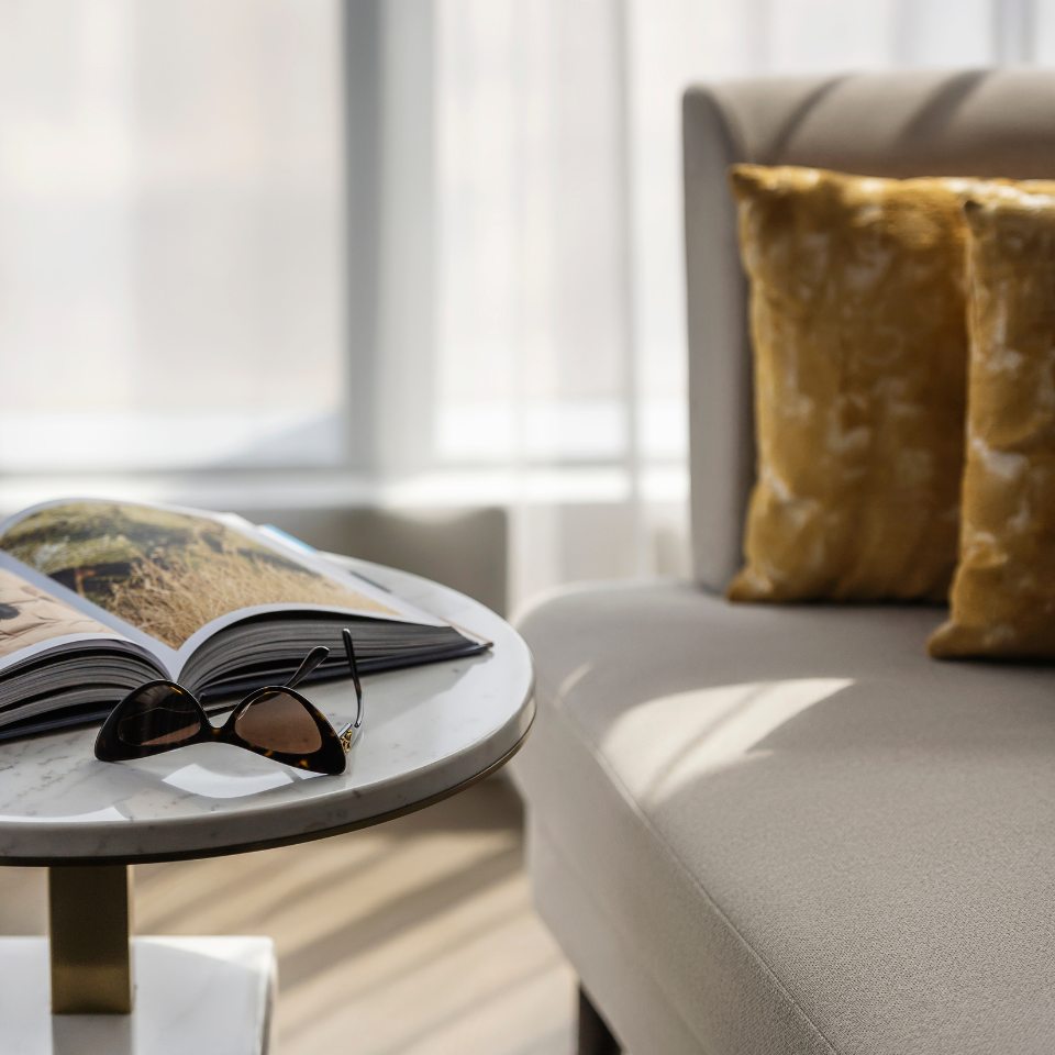 Cozy room corner with a beige sofa, two gold pillows, a marble side table holding an open book and sunglasses.