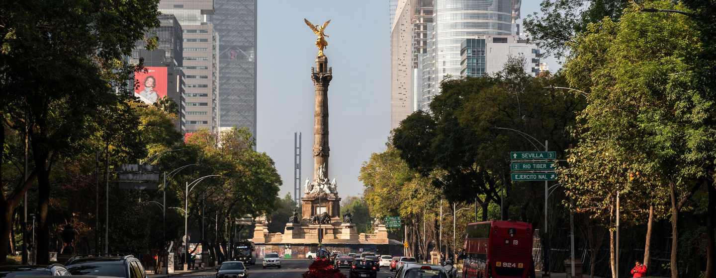 Busy city street with cars and bus, framed by trees, leading to a tall monument with a golden angel statue on top.