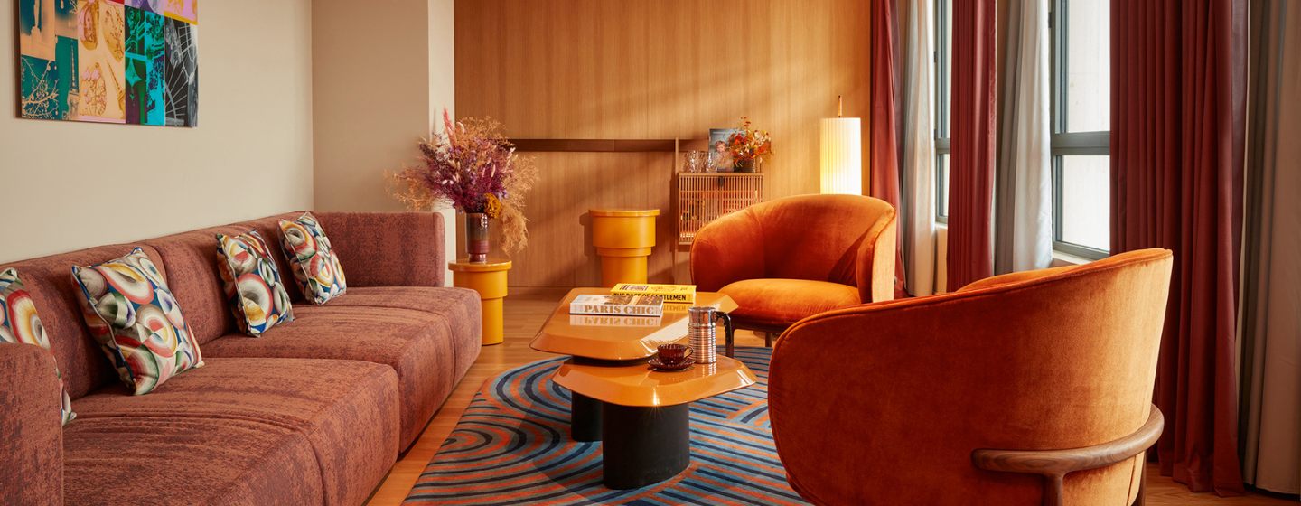 Cozy living room with warm tones; brown sofa adorned with patterned cushions, orange velvet armchairs, abstract rug, vibrant artwork, and soft lighting.
