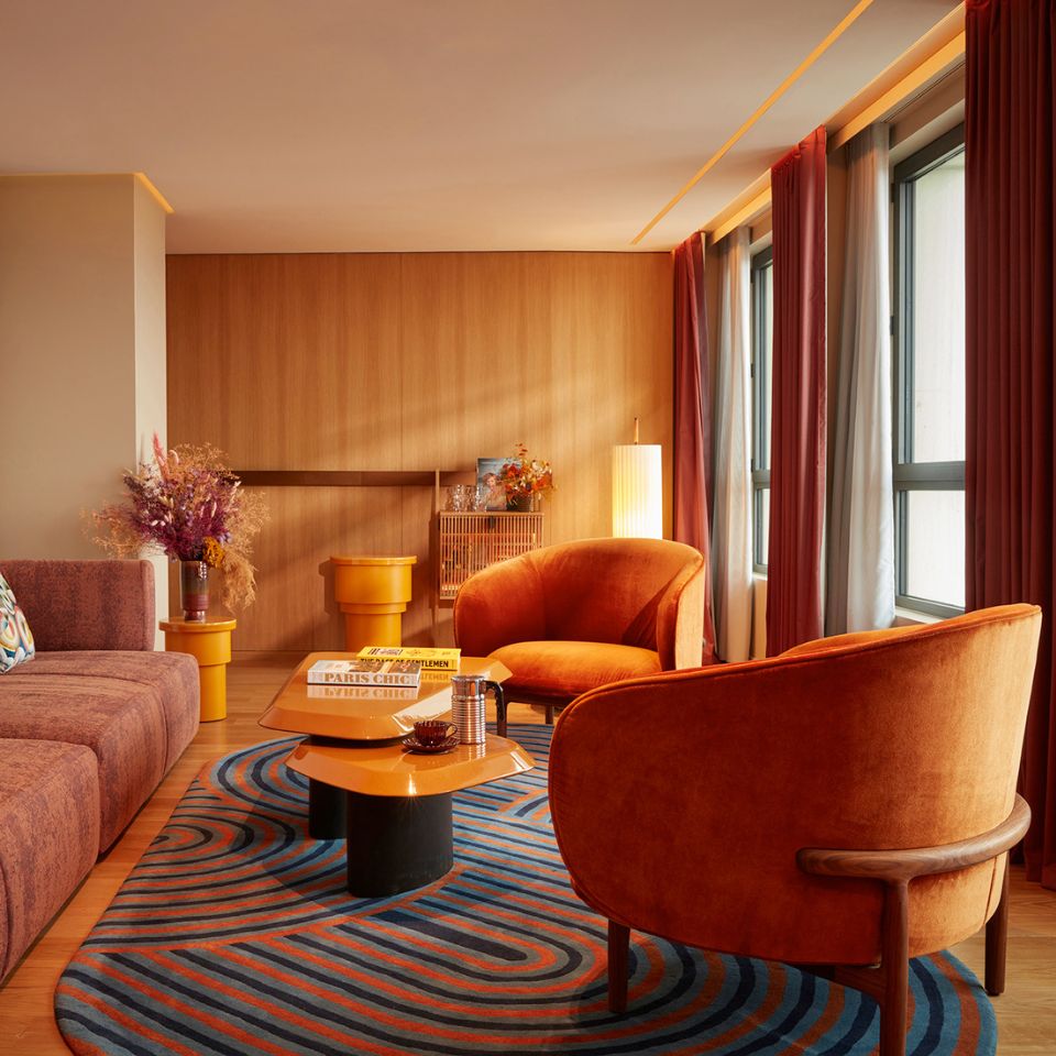 Cozy living room with warm tones; brown sofa adorned with patterned cushions, orange velvet armchairs, abstract rug, vibrant artwork, and soft lighting.