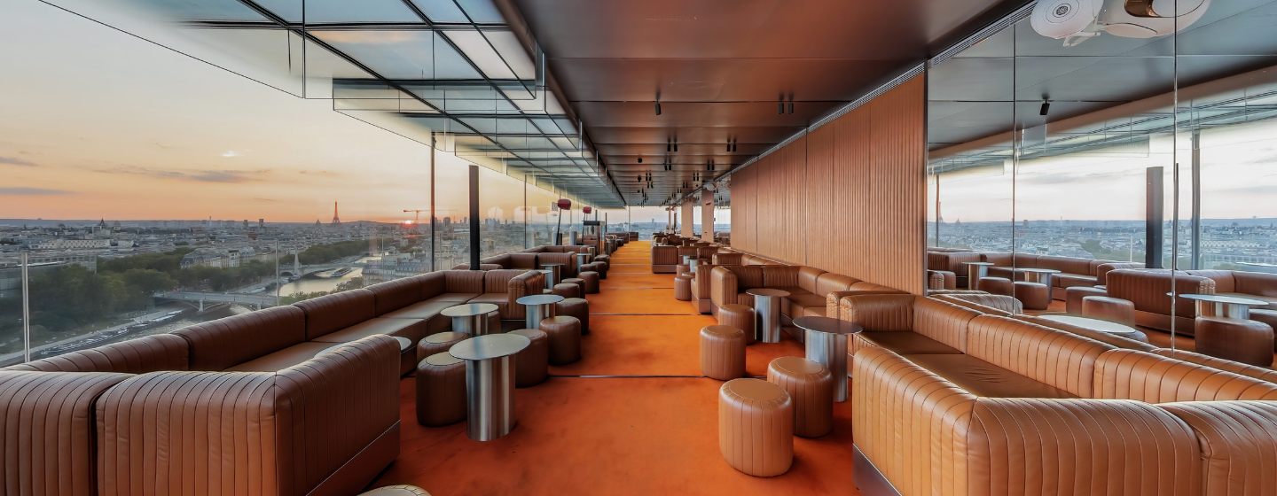 Luxurious rooftop lounge with brown leather seating and modern tables. Floor-to-ceiling windows reveal a panoramic cityscape under a serene sunset sky.