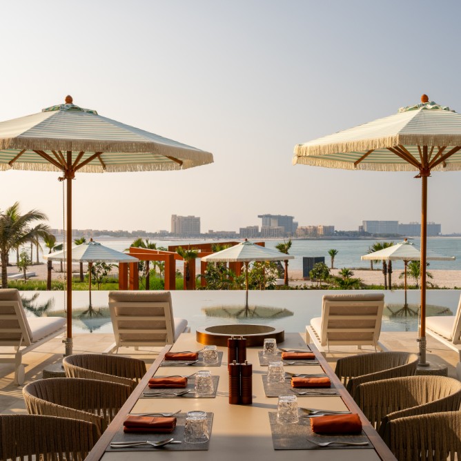 A serene beachfront dining area with a table set for six, white umbrellas, and lounge chairs. Overlooks a pool and ocean under a clear, sunny sky.