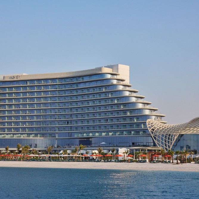 Modern, curved hotel with blue-tinted glass windows beside a sandy beach and clear ocean. Elegant and inviting under a clear blue sky.