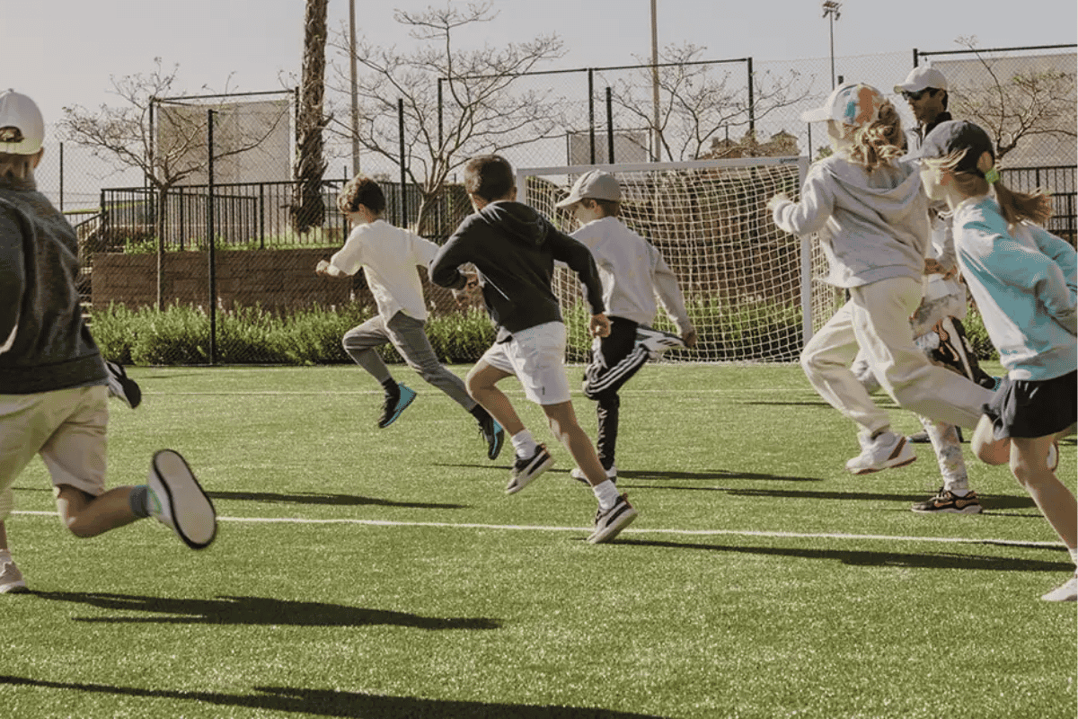Children running and playing on a sunny outdoor soccer field with a goal and fenced background.
