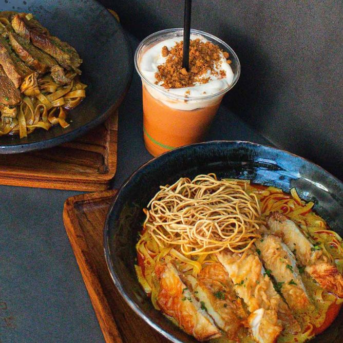 A bowl of noodle soup with fried noodles and fish fillets, a plate of fried vegetables with noodles, and a creamy iced drink topped with crumbs.