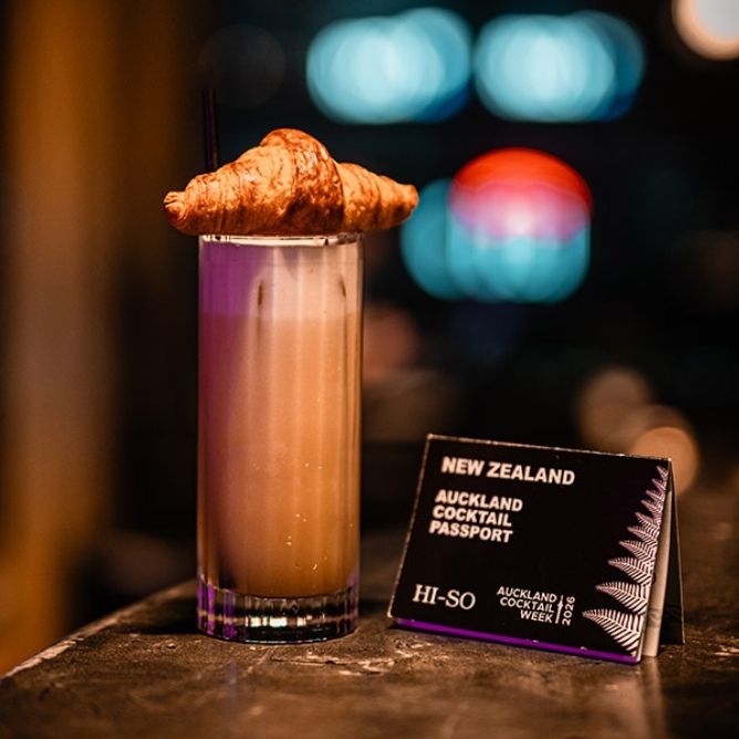 Tall creamy cocktail with a croissant garnish on top, placed next to a New Zealand Auckland Cocktail Passport card.