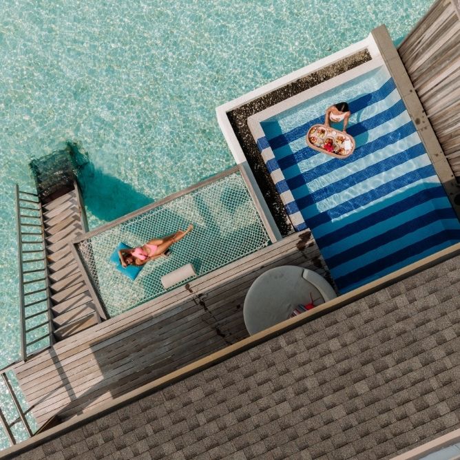 Overhead view of a woman relaxing on a net above clear water and another person with a floating tray of food in a pool next to a wooden deck.