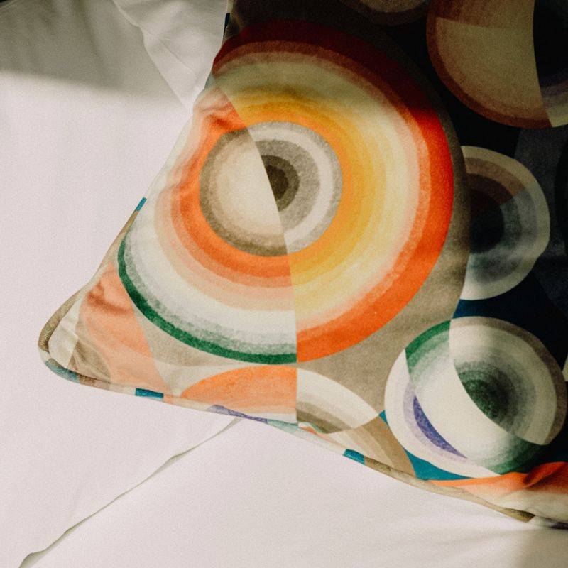 A vibrant, colorful pillow rests on a neatly made bed with a crisp white sheet.