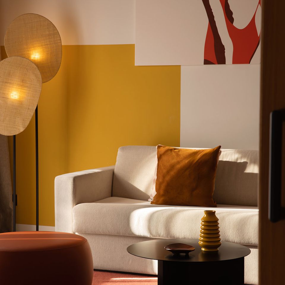 Cozy living room with beige sofa, orange pillow, round black coffee table, modern standing lamps, and mustard-yellow accent wall with artwork.