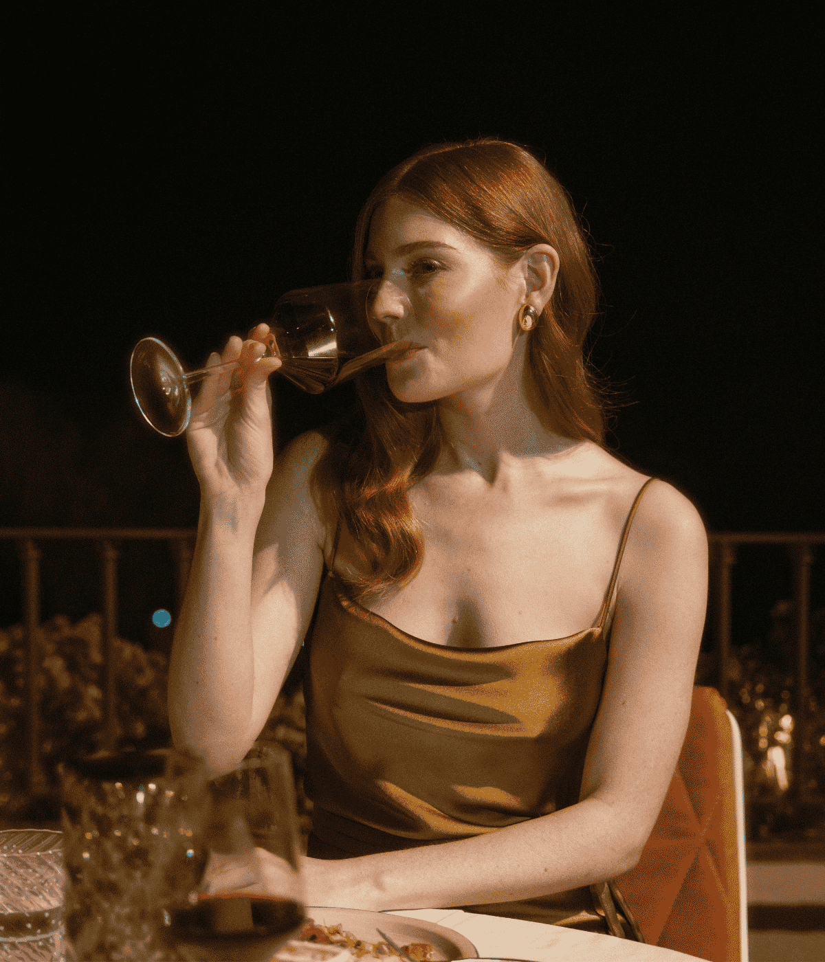 A woman in a gold dress enjoys a glass of wine while seated at a table, exuding elegance and sophistication.