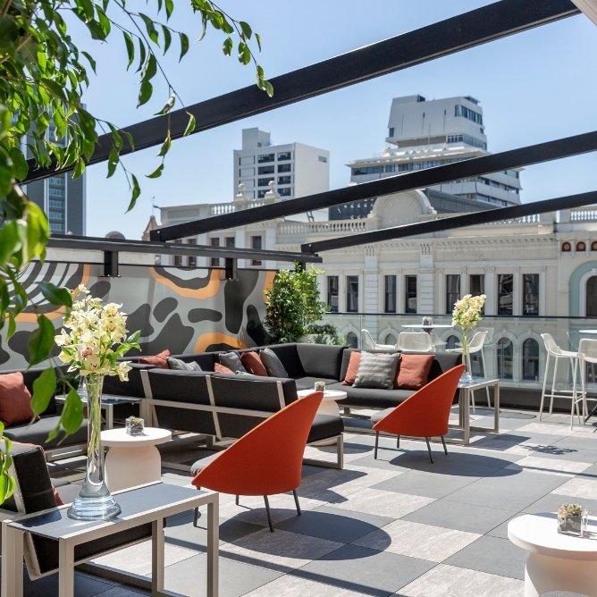 Rooftop terrace with modern seating, orange chairs, floral arrangements, and city buildings under clear blue sky.