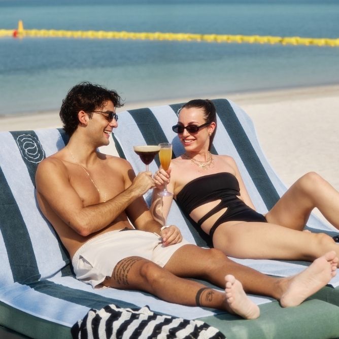 A couple wearing sunglasses lounges on beach chairs by the ocean, clinking cocktail glasses and smiling under the sun.