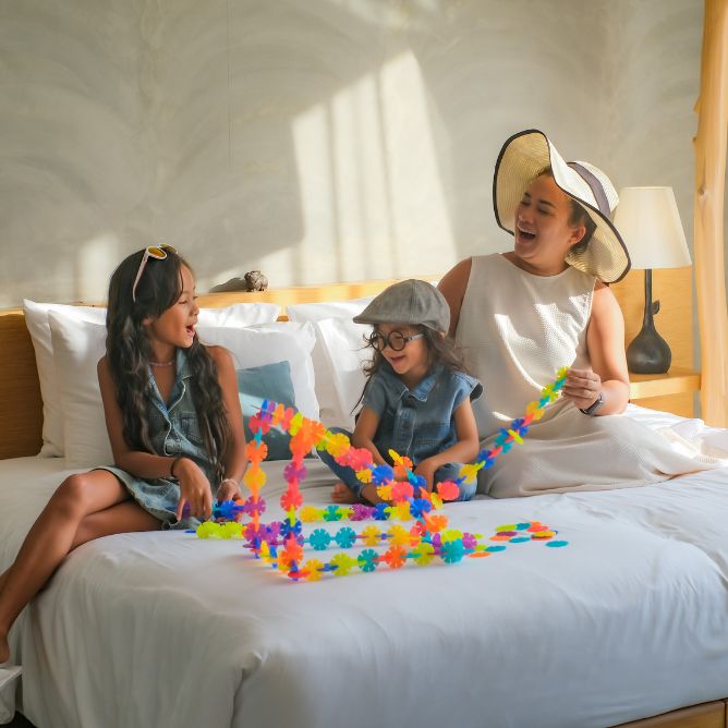 A mother in a hat laughs while playing with her two young daughters on a large white bed, surrounded by colorful plastic building blocks.