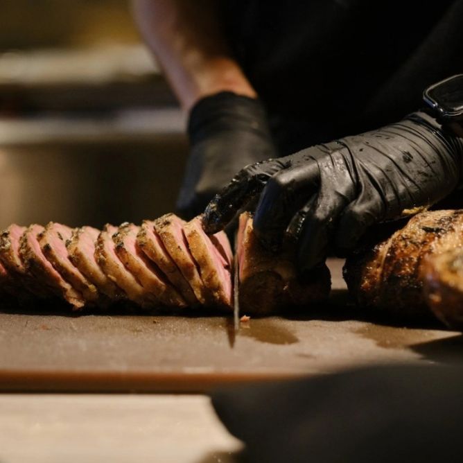 Close-up of a chef wearing black gloves slicing a tender piece of medium-rare roast meat on a cutting board.