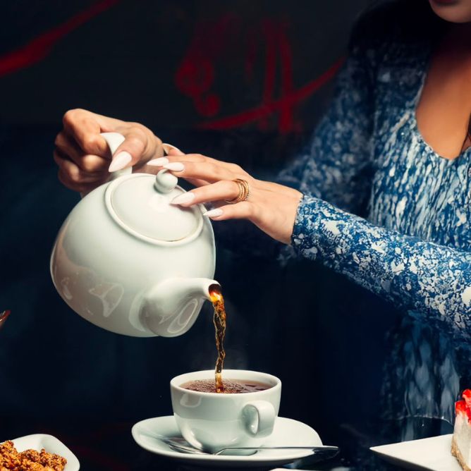 A woman gracefully pours tea from a kettle into a teapot, showcasing a moment of tea preparation.