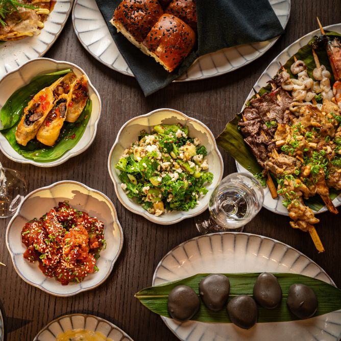 An overhead view of assorted dishes including skewers, bread rolls, salad, marinated meat, and stuffed vegetables, on a dark wooden table.