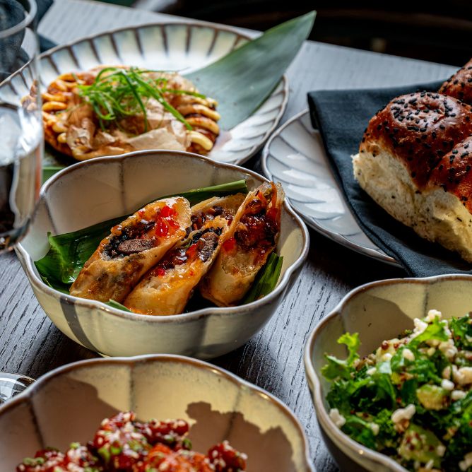 A table with bowls of various dishes, including spring rolls, salad, bread, and other assorted appetizers, arranged on a dark wooden table.