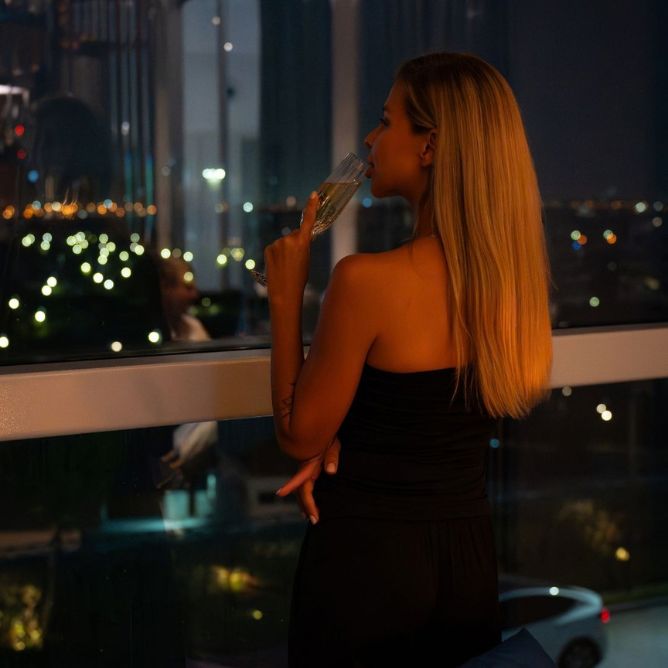 Woman enjoying champagne by a hotel window with a sparkling city night view.