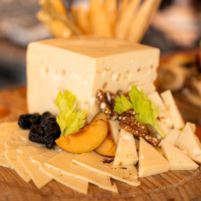 Gourmet cheese board with a large cheese block, sliced cheeses, dried plums, peaches, walnuts, and celery garnish.