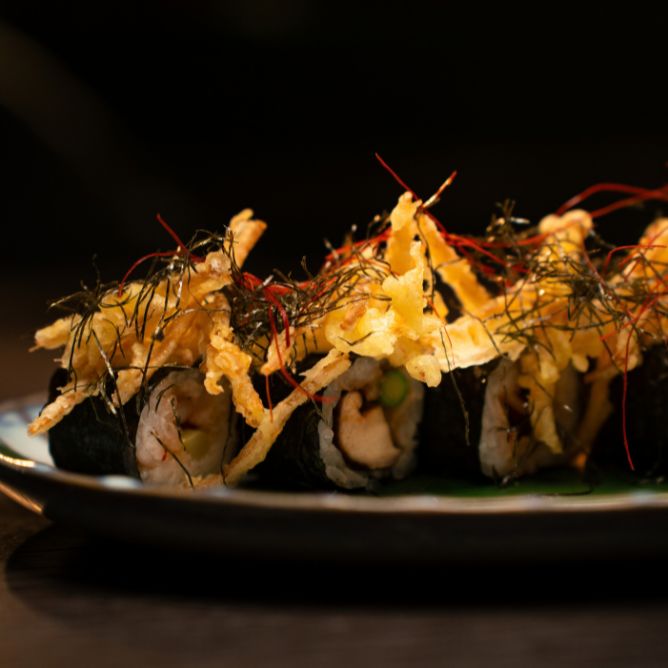 Modern fusion sushi rolls topped with crispy tempura shreds and delicate herb garnishes on a dark platter.