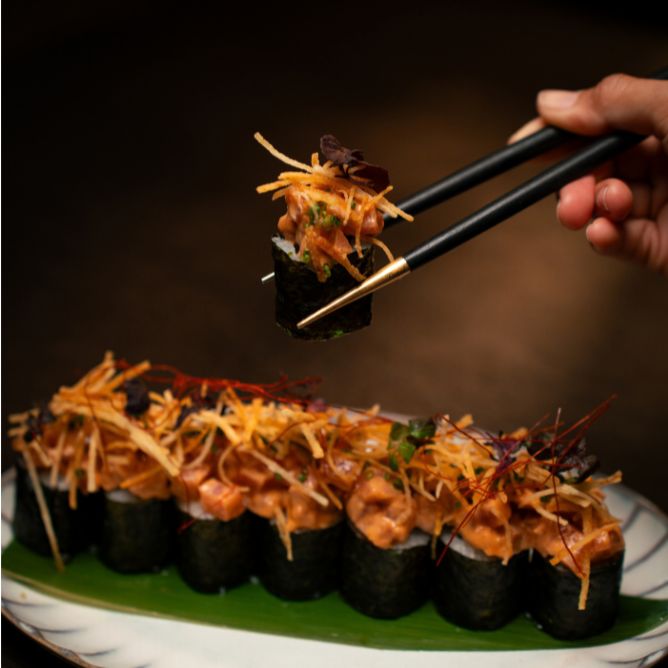 Chopsticks lifting a spicy tuna sushi roll topped with crispy shoestring potatoes from a platter on a banana leaf.