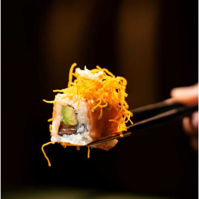 Close-up of a salmon avocado sushi roll garnished with bright orange crispy tempura flakes held by chopsticks.