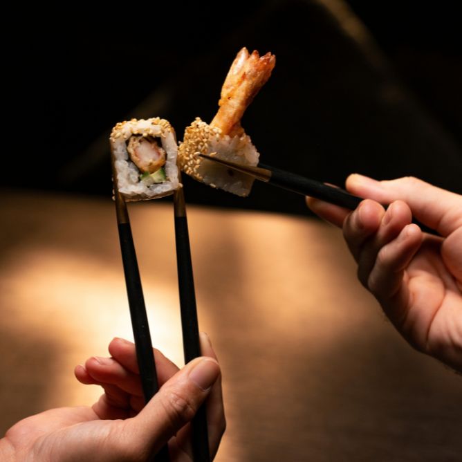 Two people toast their chopsticks together holding tempura shrimp sushi rolls at a Japanese restaurant.