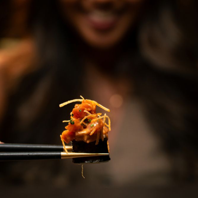Chopsticks lifting a spicy tuna sushi roll topped with crispy shoestring potatoes from a platter on a banana leaf.