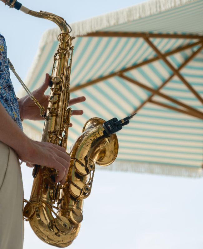 Close-up of a musician playing a golden saxophone outdoors under a blue and white striped parasol, perfect for summer live music events.