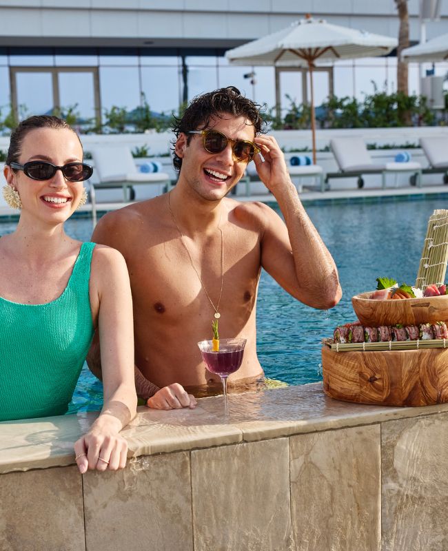 A man and woman enjoy a meal by a pool, surrounded by greenery and sunlight, creating a relaxed atmosphere.