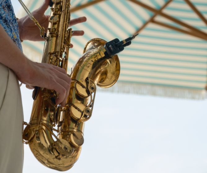 Close-up of a musician's hands playing a golden saxophone at an outdoor venue with a striped beach umbrella in the background.