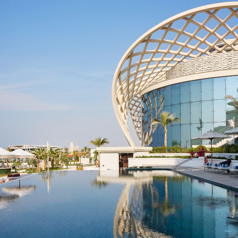 Luxury infinity pool reflecting a modern glass building with a striking white curved lattice dome structure.