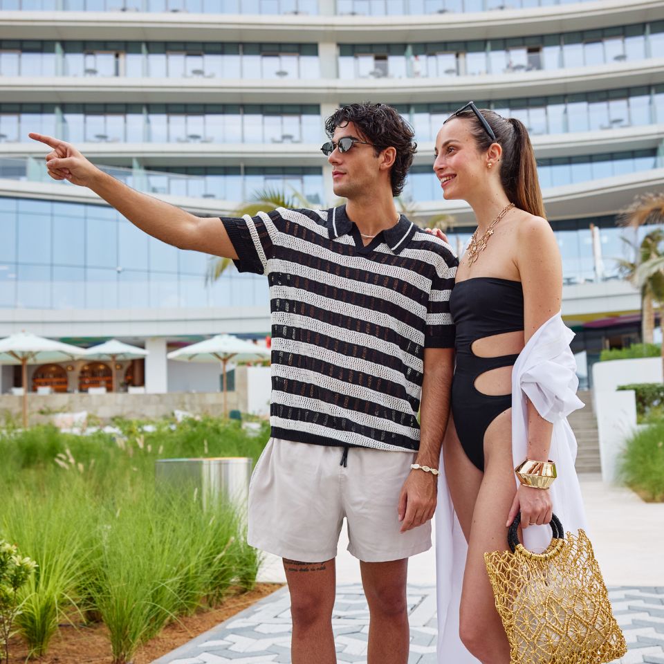 A man and woman in swimsuits pose together in front of a hotel, showcasing a sunny vacation atmosphere.