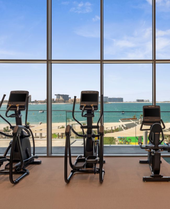 Three modern elliptical cross trainers set up in a row inside a high-end gym, positioned directly in front of floor-to-ceiling glass windows overlooking a calm turquoise sea and beach.