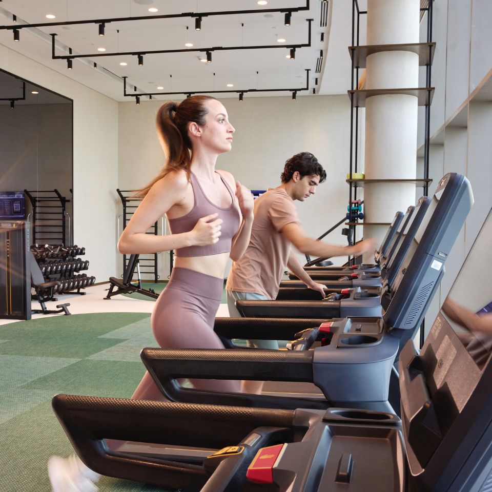 A woman in mauve athletic wear and a man in a t-shirt running side-by-side on modern treadmills in a bright, fully equipped hotel gym.