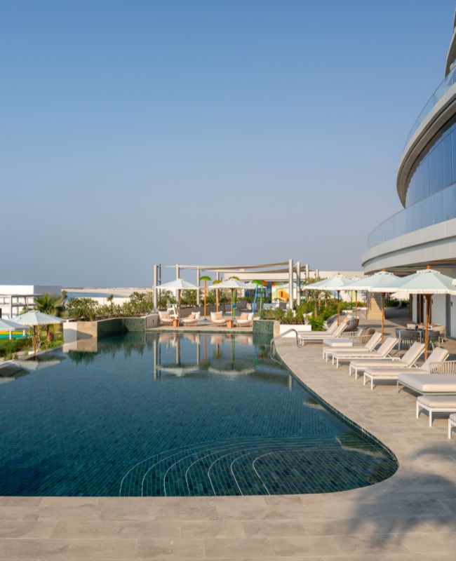 A luxurious curved infinity swimming pool with dark blue tiles, surrounded by sun loungers and modern resort architecture, offering a panoramic view of the horizon.