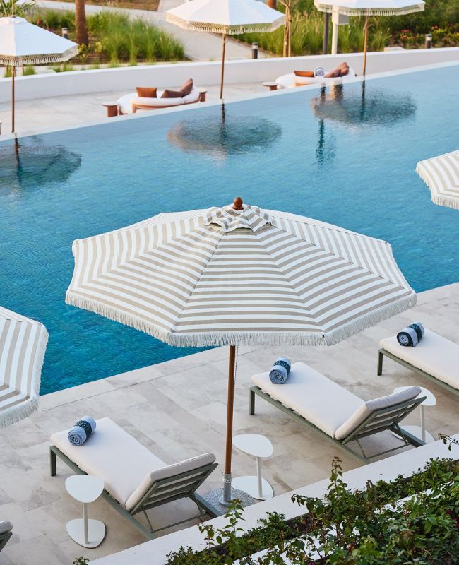 An elegant resort swimming pool area with pristine turquoise water, lined with white sun loungers and stylish beige-and-white striped parasols featuring fringed edges.