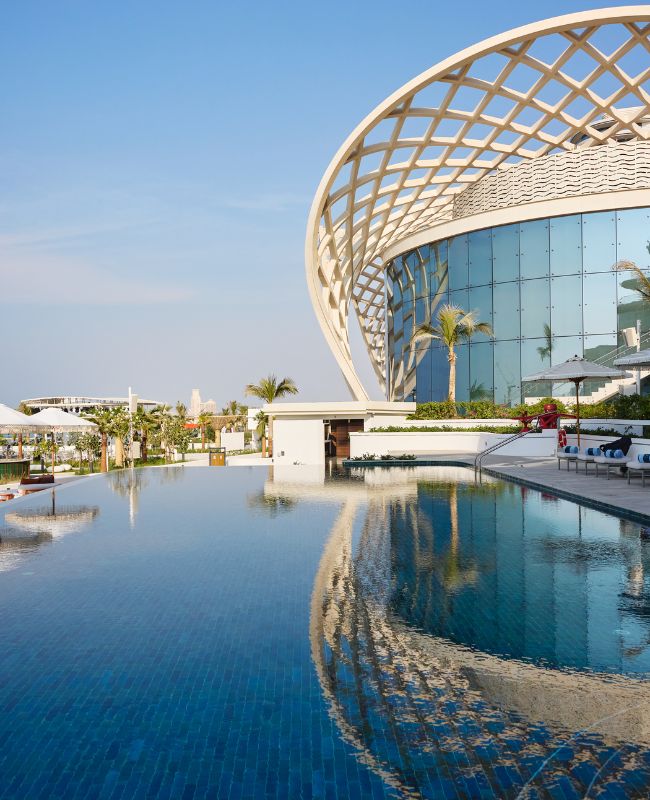 A luxurious infinity-edge resort pool reflecting a modern building with a glass facade and a massive, iconic curved wooden lattice arch structure under a clear blue sky.