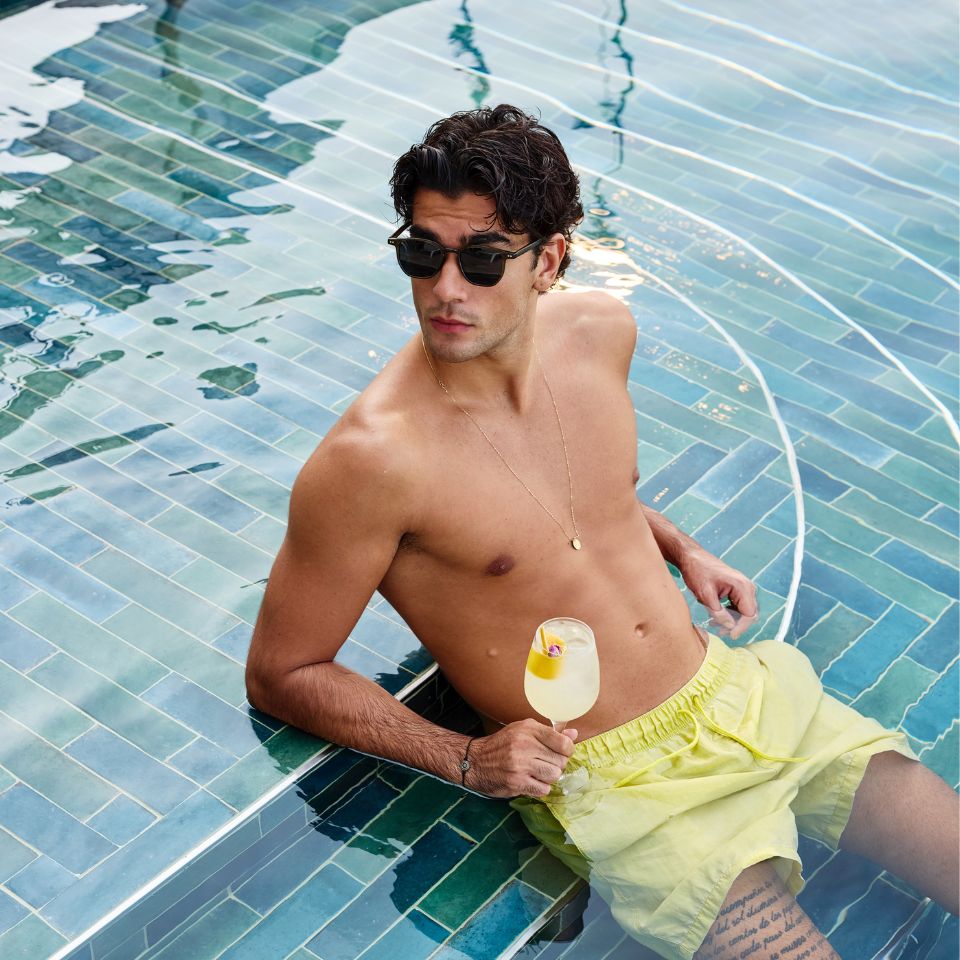 A young man in yellow swim trunks and sunglasses relaxing in a clear blue tiled swimming pool, holding a cold citrus cocktail during a sunny day.