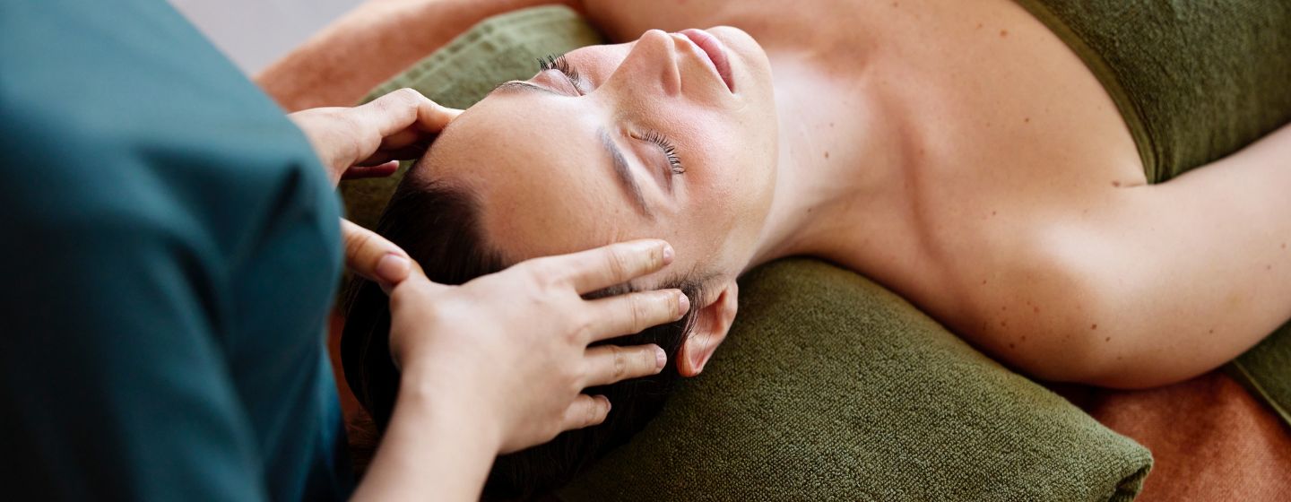 Panoramic close-up of a relaxed woman lying down with eyes closed, receiving a soothing head and temple massage from a professional spa therapist.