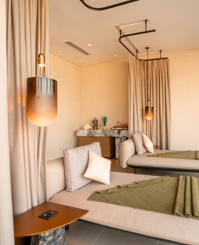 Peaceful spa relaxation room featuring two plush daybeds with green throws, separated by flowing privacy curtains and illuminated by glowing amber glass pendant lamps.