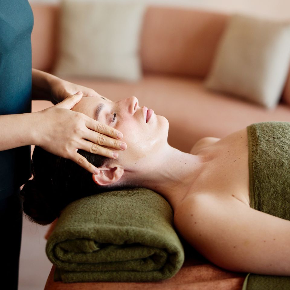 Close-up of a relaxed woman lying down with eyes closed, receiving a soothing facial and temple massage from a professional spa therapist.