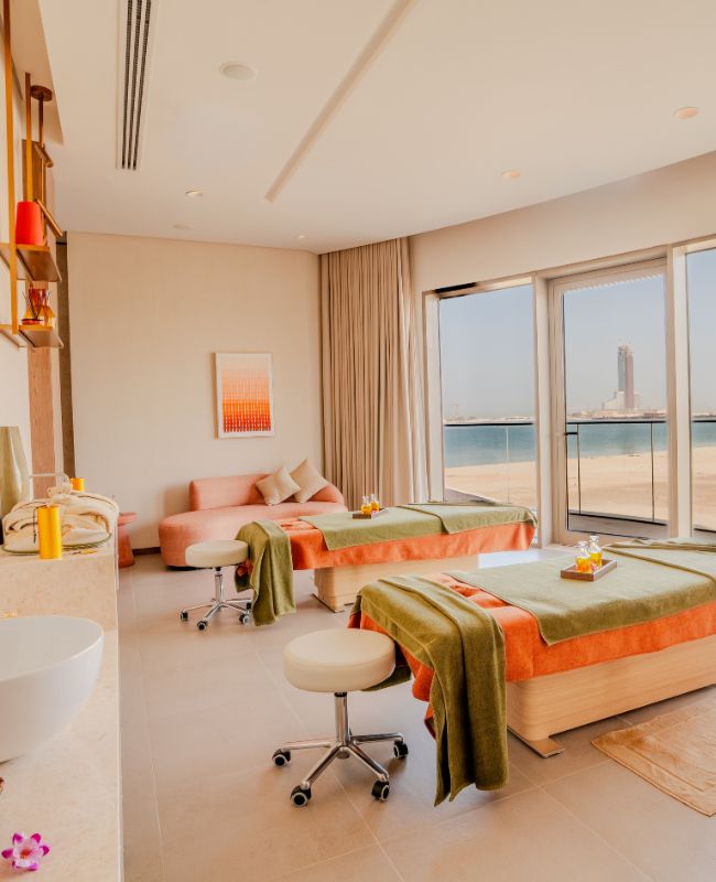 A luxurious couple's spa treatment suite featuring two massage tables with green covers, a plush pink lounge sofa, and large sliding glass doors revealing a pristine sandy beach and turquoise sea.