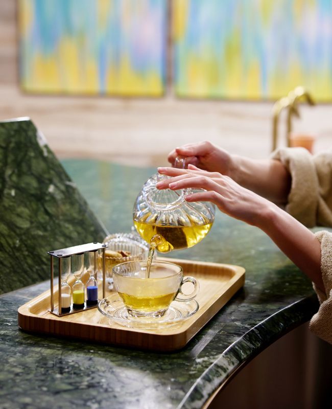 Close-up of a spa guest in a bathrobe pouring fresh herbal tea from a clear glass teapot into a matching teacup on a wooden tray with a decorative sand timer.
