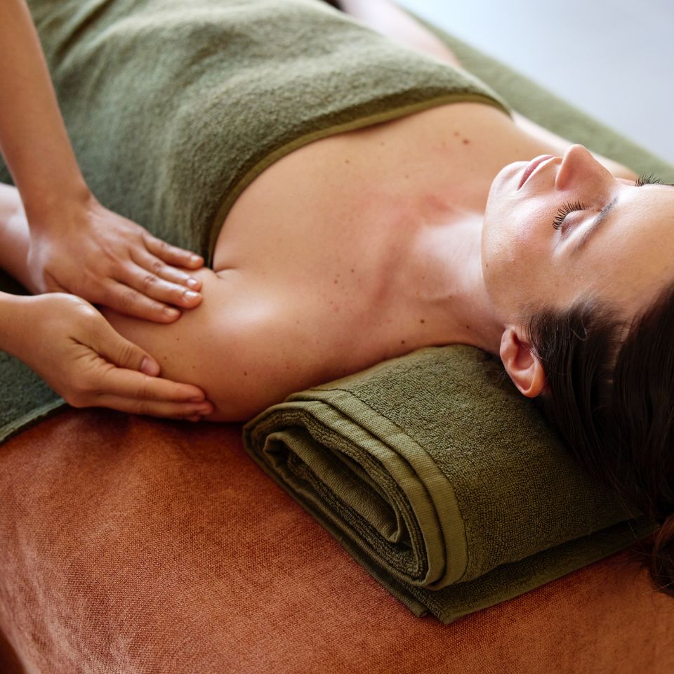 A close-up, panoramic shot of a relaxed woman lying on a massage table with eyes closed, receiving a therapeutic shoulder and neck massage from a professional therapist.