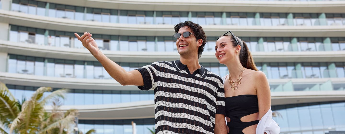 A happy young couple in stylish summer wear smiling as the man points towards the distance in front of a modern hotel building with curved balconies.