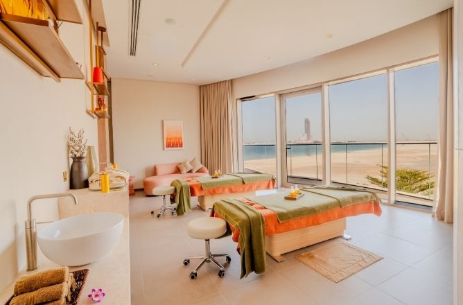 http://A%20bright,%20spacious%20couple's%20massage%20room%20featuring%20two%20treatment%20beds,%20a%20lounge%20sofa,%20a%20vanity%20sink,%20and%20large%20glass%20sliding%20doors%20looking%20out%20to%20a%20sandy%20beach%20and%20ocean.