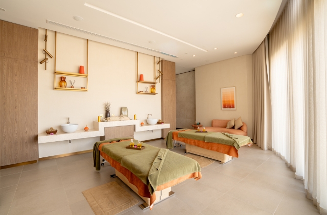 http://A%20luxurious%20couple's%20spa%20treatment%20room%20featuring%20two%20neatly%20made%20massage%20tables%20with%20olive%20green%20blankets,%20dual%20vanity%20sinks,%20golden%20decorative%20shelving,%20and%20a%20plush%20daybed%20in%20the%20corner.